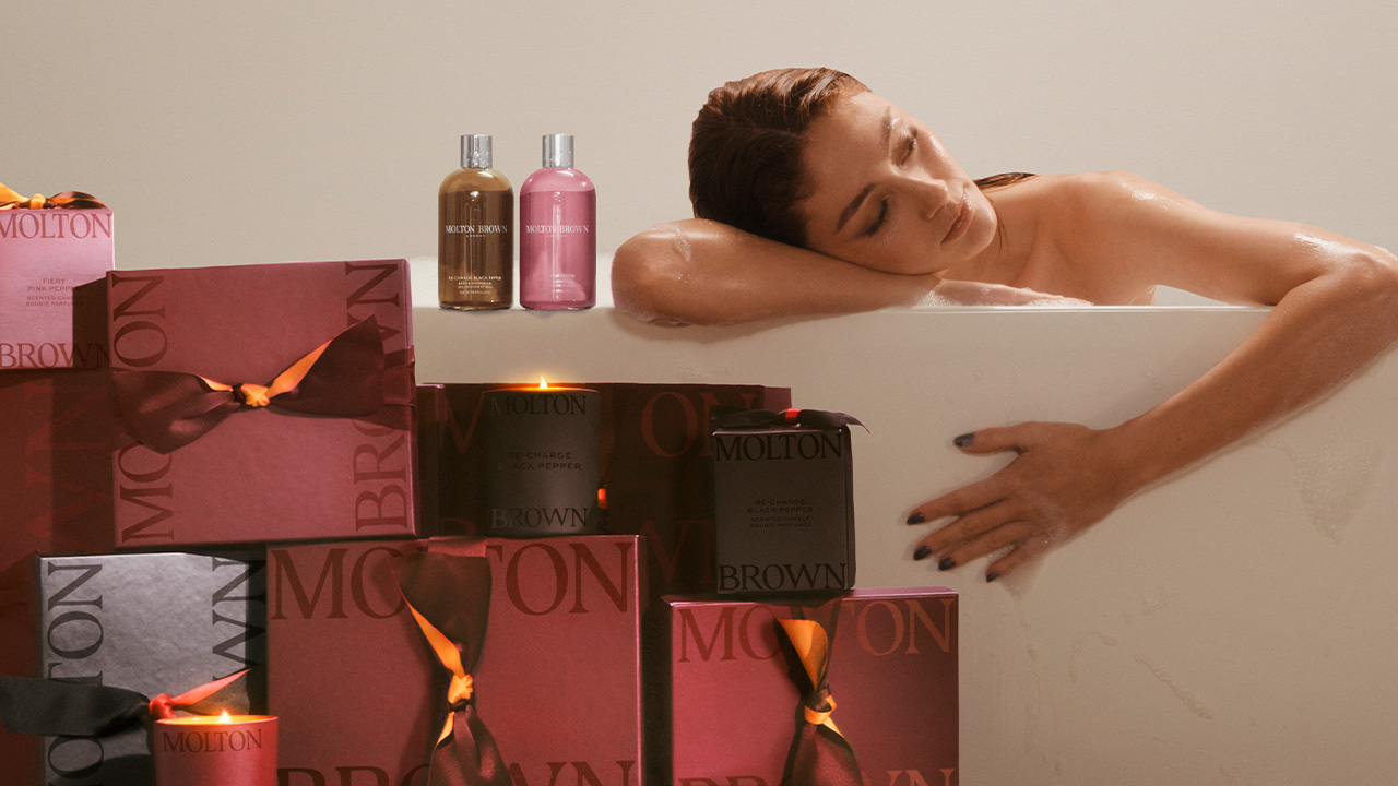 A relaxed person is resting in a bathtub, surrounded by neatly stacked luxurious gift boxes tied with orange and brown ribbons. Two bottles of bath products are placed at the edge of the tub. The scene conveys a sense of calm and indulgence.